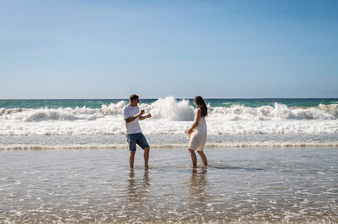 Couple playing in shallow waves on a Cornwall beach with a bright blue sky and crashing surf in the background.