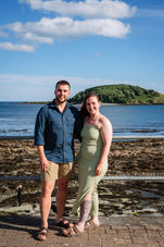 Couple standing near the shoreline with Looe Island and clear blue skies in the background.