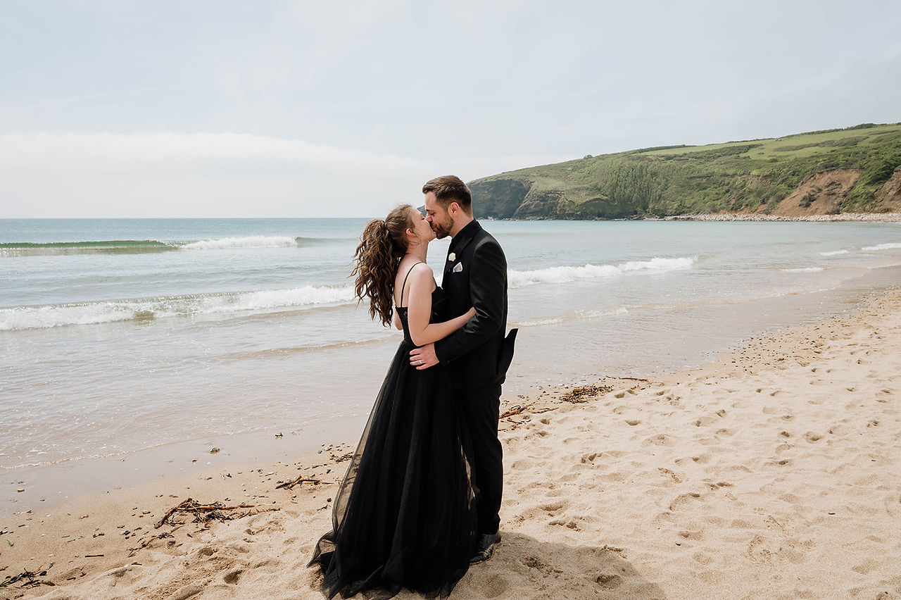 Elopement couple kissing on the beach at Praa Sands, Cornwall, with rolling waves and a picturesque coastal backdrop