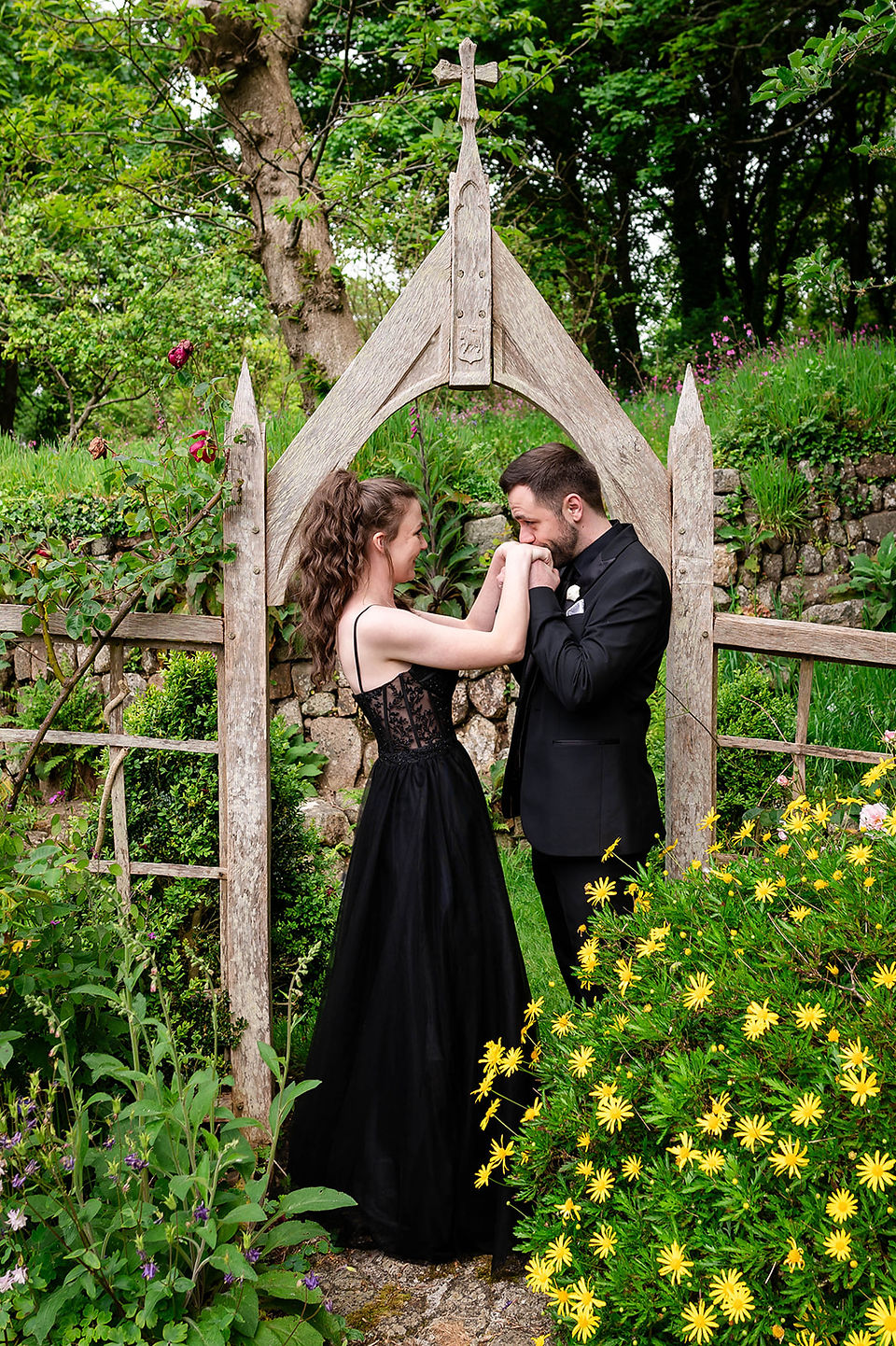 Elopement couple sharing a tender moment under a wooden archway in the gardens of Pengersick Castle, Cornwall, surrounded by greenery and yellow flowers