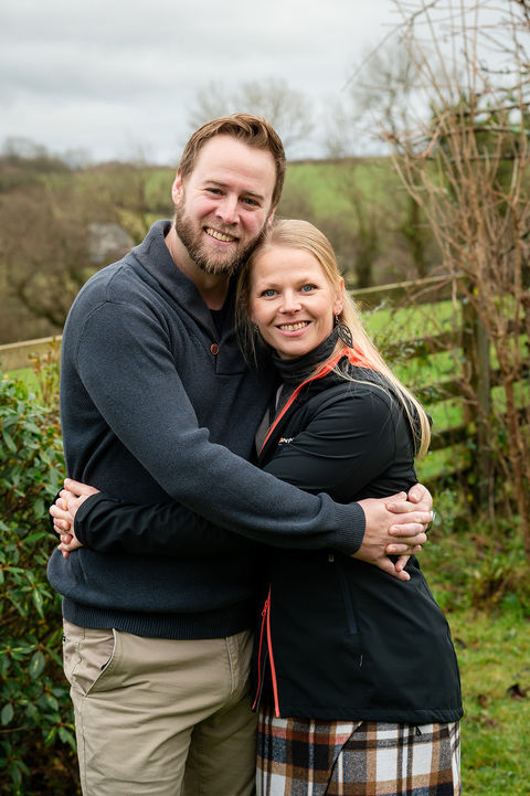 Couple smiling and embracing outdoors in a peaceful countryside setting with green fields and trees in the background.