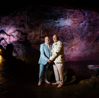 Two grooms holding hands at their Wookey Hole wedding in Somerset, standing in a beautifully lit cave with colorful rock formations and dramatic lighting.