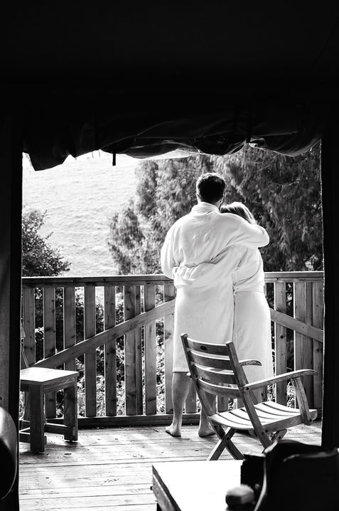 Couple in robes standing on a wooden balcony, embracing and looking out at a serene countryside view in black and white