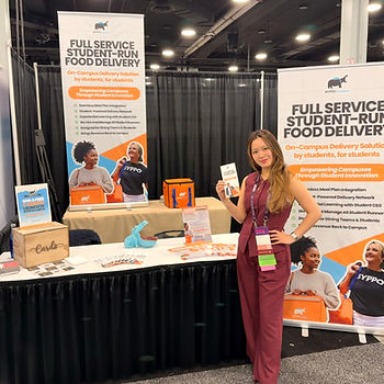Victoria Liu stands smiling at a booth with "Full Service Student-Run Food Delivery" banners, holding a brochure. Background features posters, an orange bag, and display materials.