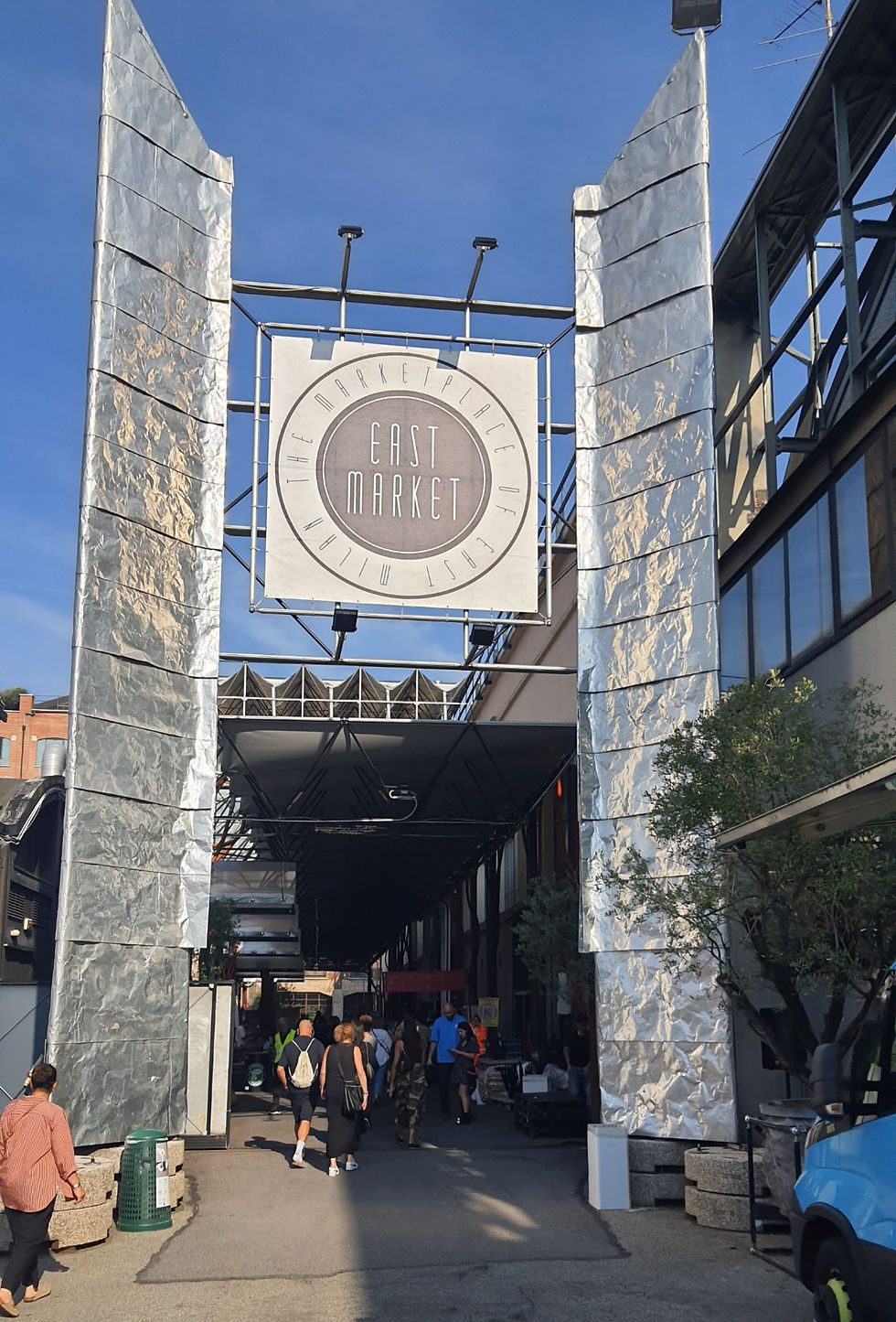 כניסה לשוק EAST MARKET עם שלט גדול, אנשים הולכים תחת קשתות כסופות בשמיים כחולים.