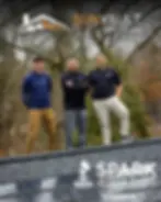 The three owners of Sun Vault Roofing stand at the Ridge of a black shingle roof above an array of the solar shingles with trees in the background.