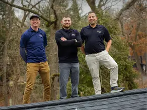 The three owners of Sun Vault Roofing stand at the Ridge of a black shingle roof above an array of the solar shingles with trees in the background.
