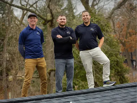 The three owners of Sun Vault Roofing stand at the Ridge of a black shingle roof above an array of the solar shingles with trees in the background.