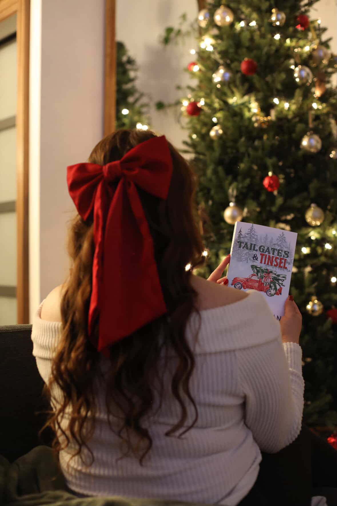 nnnnPhotograph of a woman wearing a cream sweater with long dark hair tied back in a red bow, holding a copy of Tailgates & Tinsel by J.C. Hannigan in front of a beautifully decorated Christmas tree.