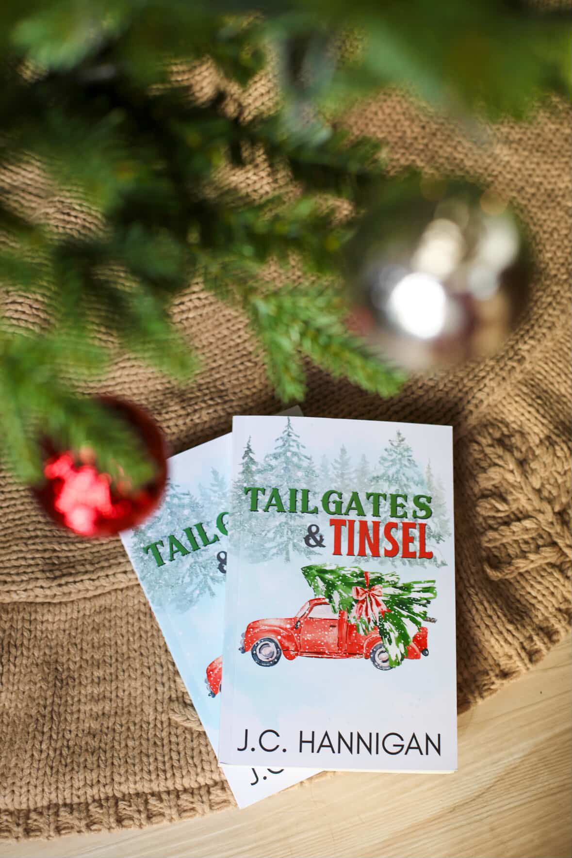 nnnnPhotograph of two copies of Tailgates & Tinsel by J.C. Hannigan underneath an out-of-focus Christmas tree.