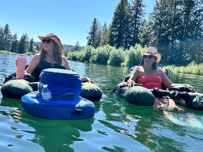 Two women floating side by side in inflatable float chairs on the Spokane River, both wearing sun hats and sunglasses as they relax on the water in bright summer light, lifestyle photo showing a calm, carefree river float outing, photographed by North Idaho Explorer photographer Carly Agnew.