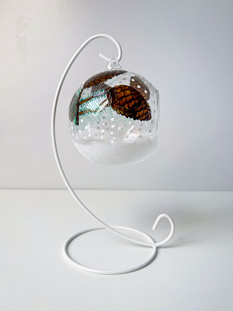 Thumbnail: Hand-painted pine cone glass candle holder with amber brown pine cones and green needles, displayed on a white metal stand.