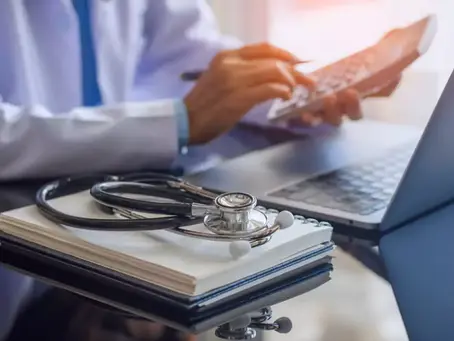 a physician using a calculator at his computer with a notebook and stethoscope on the table. Image meant to symbolize the financial synergies in healthcare M&A