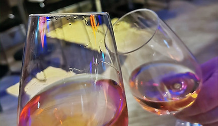2 Weingläser die in extremer Nahaufnahme anstoßen. im Hintergrund eine Bar mit Stühlen.