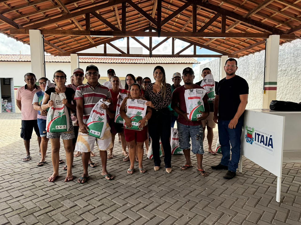 Grupo de moradores reunidos em espaço coberto recebendo sacos de sementes, ao lado de representantes locais, com identificação da Prefeitura de Itajá ao fundo.