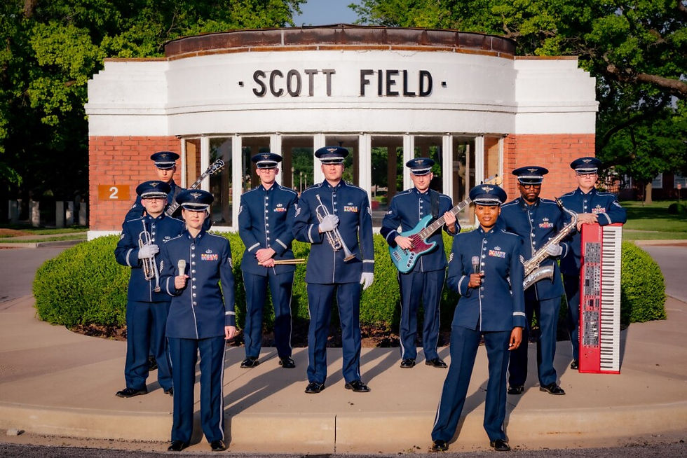 Grupo de músicos da Força Aérea dos Estados Unidos posa uniformizado em frente ao prédio Scott Field. Os integrantes seguram instrumentos como trompete, guitarra, saxofone e teclado durante registro oficial ao ar livre.