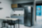 Cozinha compacta e moderna com mesa de duas cadeiras, armários cinza e detalhes em madeira. O ambiente possui geladeira inox, micro-ondas, pia e iluminação clara, com corredor ao fundo levando para outros cômodos.