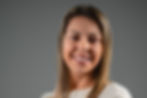 Retrato de uma mulher sorrindo para a câmera, com os braços cruzados, vestindo uma blusa branca de mangas longas. A foto foi feita diante de um fundo cinza liso, com iluminação suave e aparência profissional.