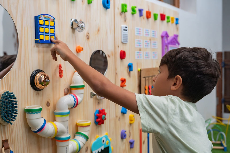Criança interage com painel sensorial de madeira equipado com jogos, objetos coloridos, espelhos e elementos educativos.