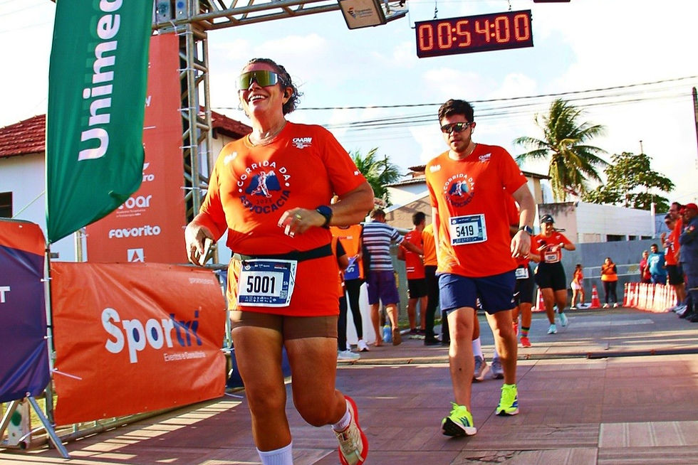 Corredores com camisetas laranja cruzam a linha de chegada de uma corrida de rua, sob pórtico com cronômetro digital.