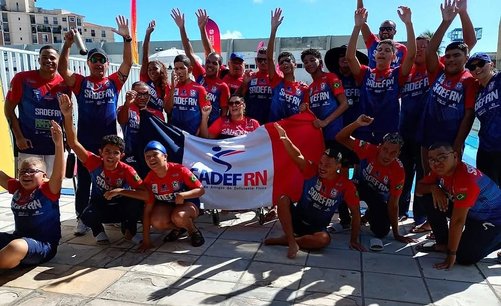 Grupo de atletas e integrantes da SADEF/RN posa ao redor de uma bandeira da instituição em área externa próxima a uma piscina. Os participantes sorriem, levantam os braços e celebram durante evento esportivo paralímpico.