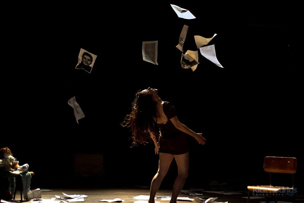 Performer em cena escura inclina o corpo para trás enquanto várias folhas de papel e fotografias caem do alto. A iluminação dramática destaca o movimento do corpo e os papéis suspensos no ar, criando atmosfera intensa e poética.