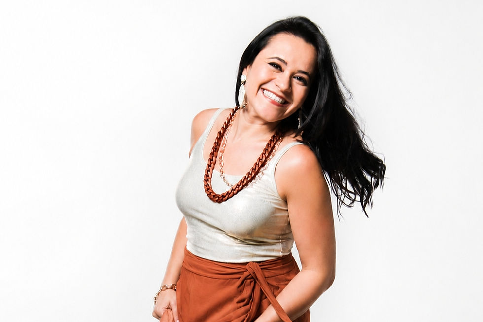 Retrato em estúdio da cantora potiguar Nara Costa sorrindo para a câmera, usando blusa clara e saia longa em tom terracota esvoaçante. A artista posa em movimento sobre fundo branco, transmitindo leveza, alegria e elegância.