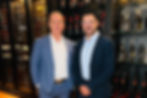 Dois homens posam lado a lado em uma adega, cercados por prateleiras repletas de garrafas de vinho e destilados. Um veste terno azul-claro e camisa branca, enquanto o outro usa blazer azul-marinho, camisa azul-clara, calça escura, tênis branco e óculos. Ambos sorriem para a câmera em um ambiente sofisticado.