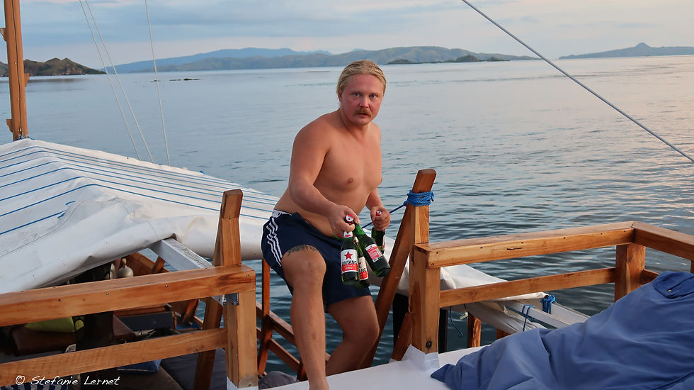 Mann mit Bierflaschen auf Boot, wirkt überrascht. Ruhiges Wasser und Hügel im Hintergrund. Blau-weiße Abdeckung auf Deck.