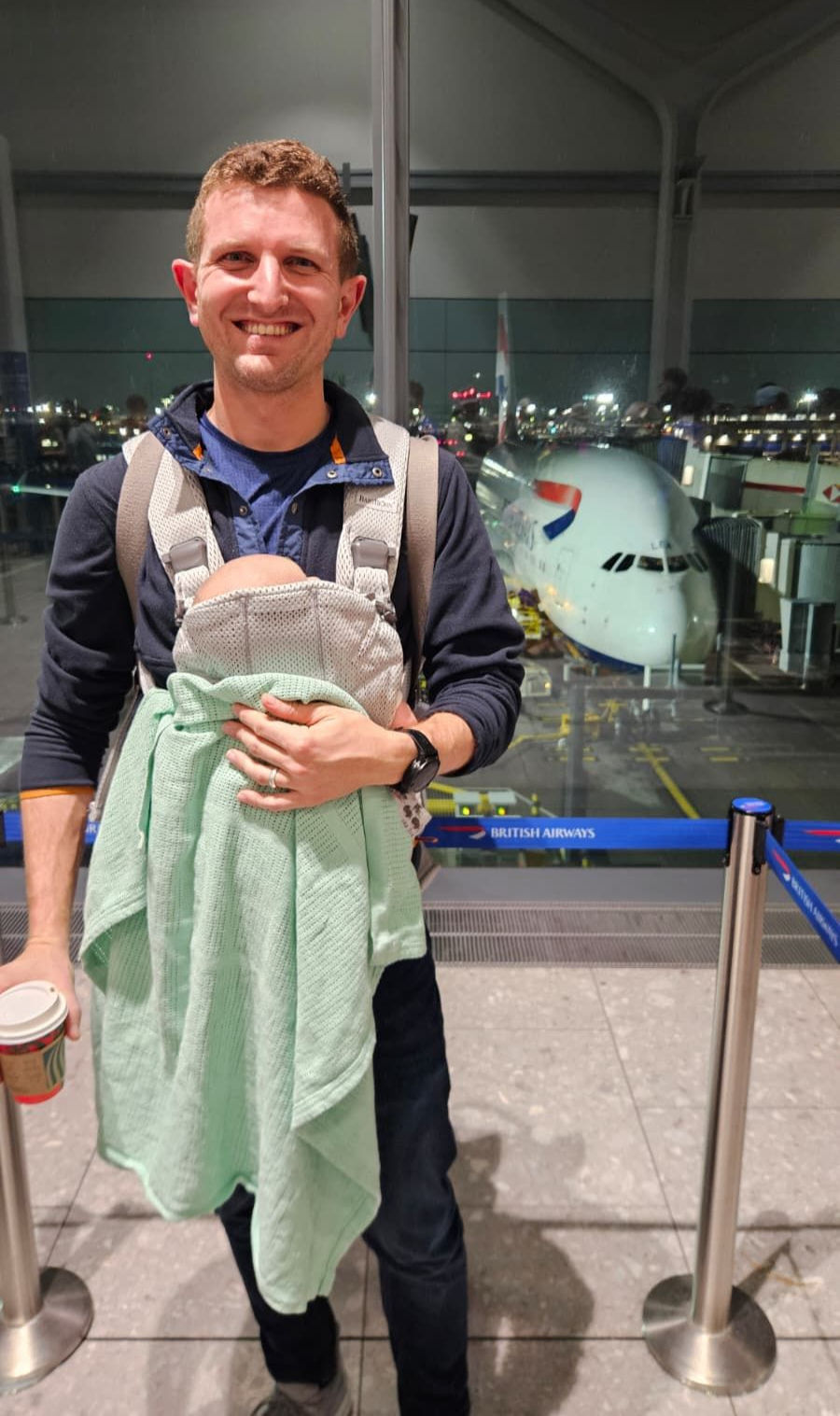 Daan van Heerden at the airport, carrying his young son in a baby carrier. A plane can be seen through the window in the background.