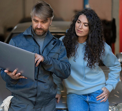 Zwei menschen beobachten einen Laptop in einer Kfz-Werkstatt während sie sich an einem Auto anlehnen.