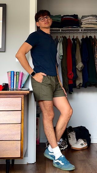 Young man standing agains a doorframe in a navy blue shirt and olive green shorts.