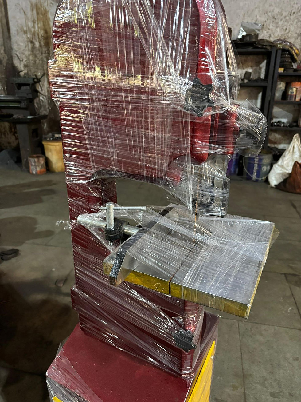 WoodWorking Bandsaw Machine Manufacturer In Rajkot, Gujarat, India. Red industrial bandsaw wrapped in transparent plastic, placed in a workshop with a concrete floor and shelves in the background.