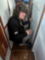 Person with curly hair kneels in a narrow hallway, smiling while unlocking a door. Wears a black jacket and gray pants. Cozy atmosphere.