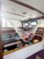 A kitchen on a boat with a hammock holding fruits and vegetables. Wooden cabinets, pantry items, a sink, and a "Fire Blanket" sign are visible.