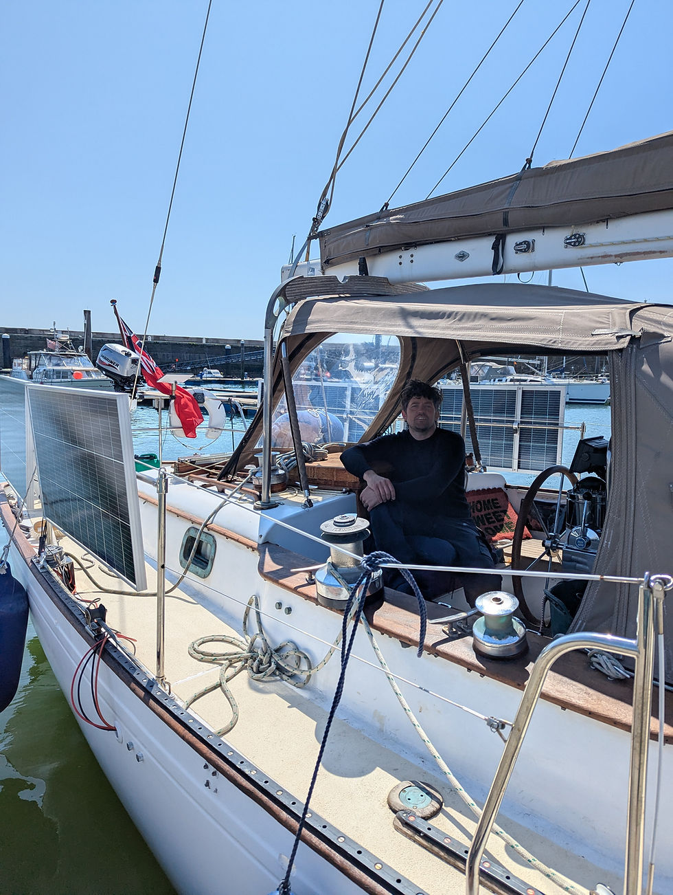 Man sitting on a sailboat docked in a sunny marina, surrounded by ropes and a solar panel. A red flag is also visible.