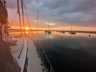 Sailboat deck at sunset with calm water, moored boats, and an orange sky. The scene is peaceful and serene.