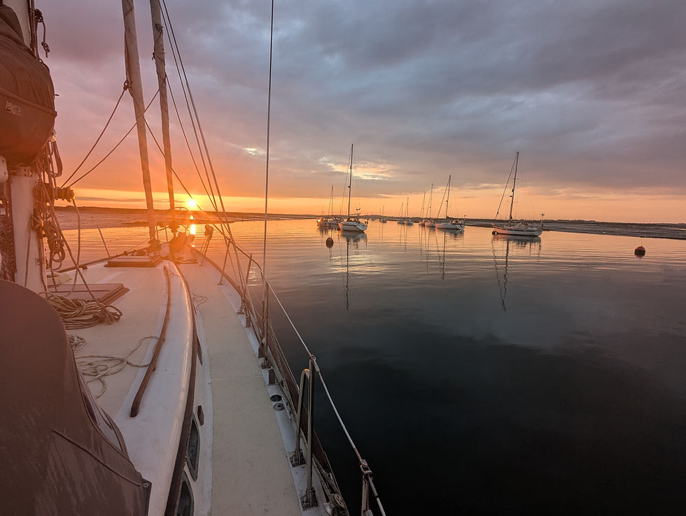Sailboat deck at sunset with calm water, moored boats, and an orange sky. The scene is peaceful and serene.