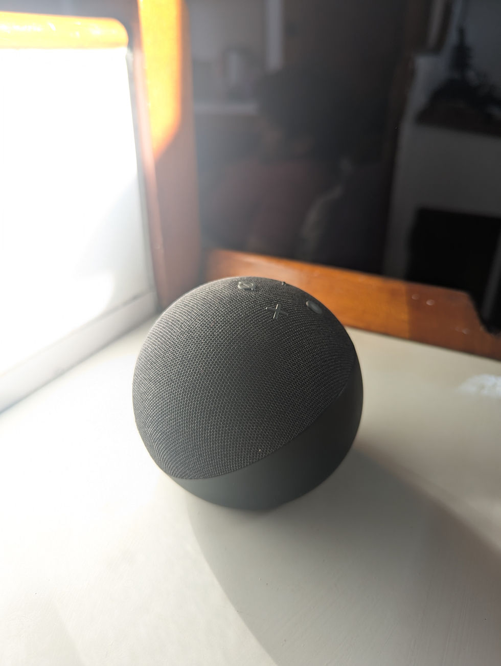 A black spherical smart speaker on a sunlit table with a shadow. A blurred background shows a person sitting, creating a relaxed ambiance.