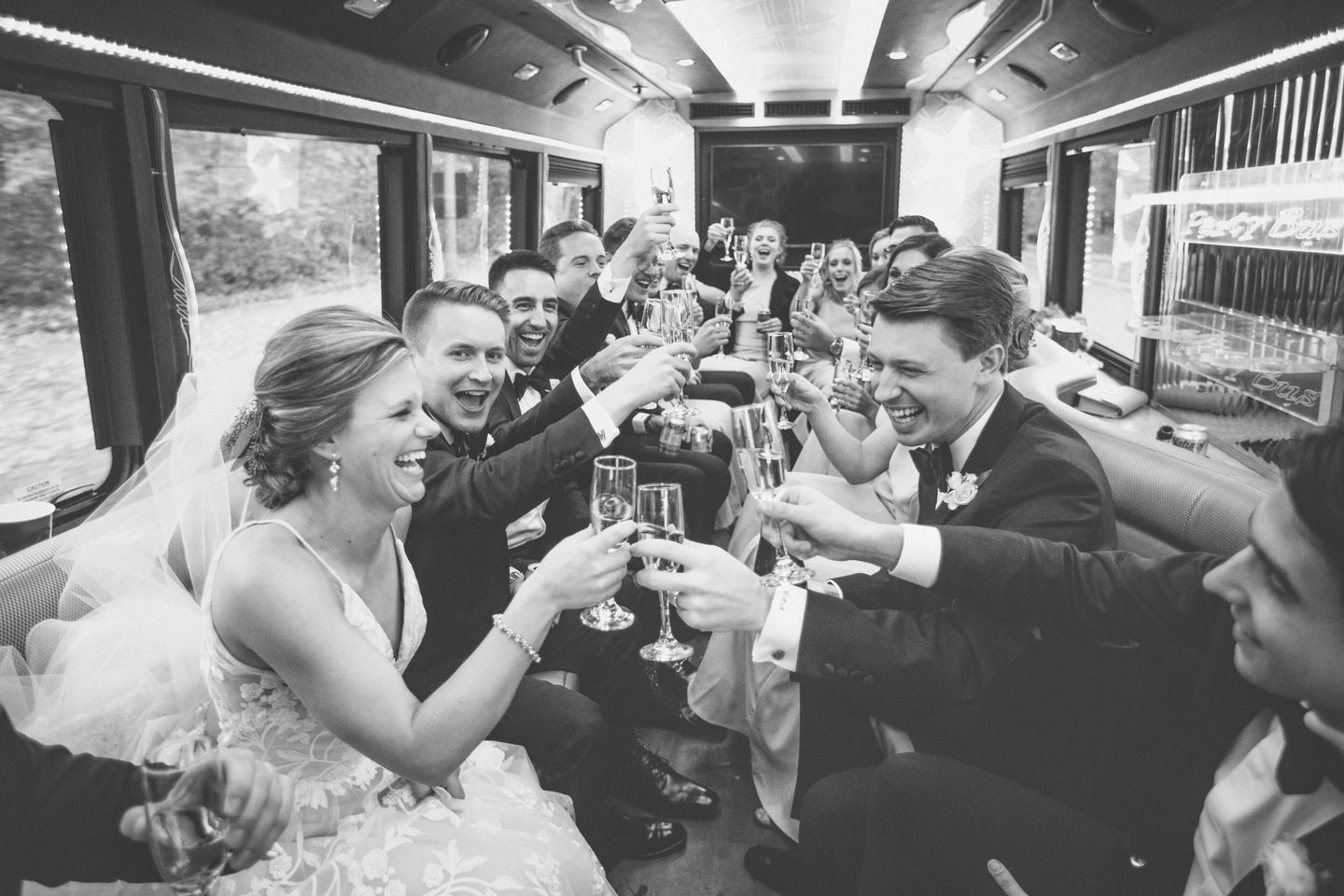 black and white candid photo bride and groom laughing and toasting champagne with a large group of friends inside a limousine 
