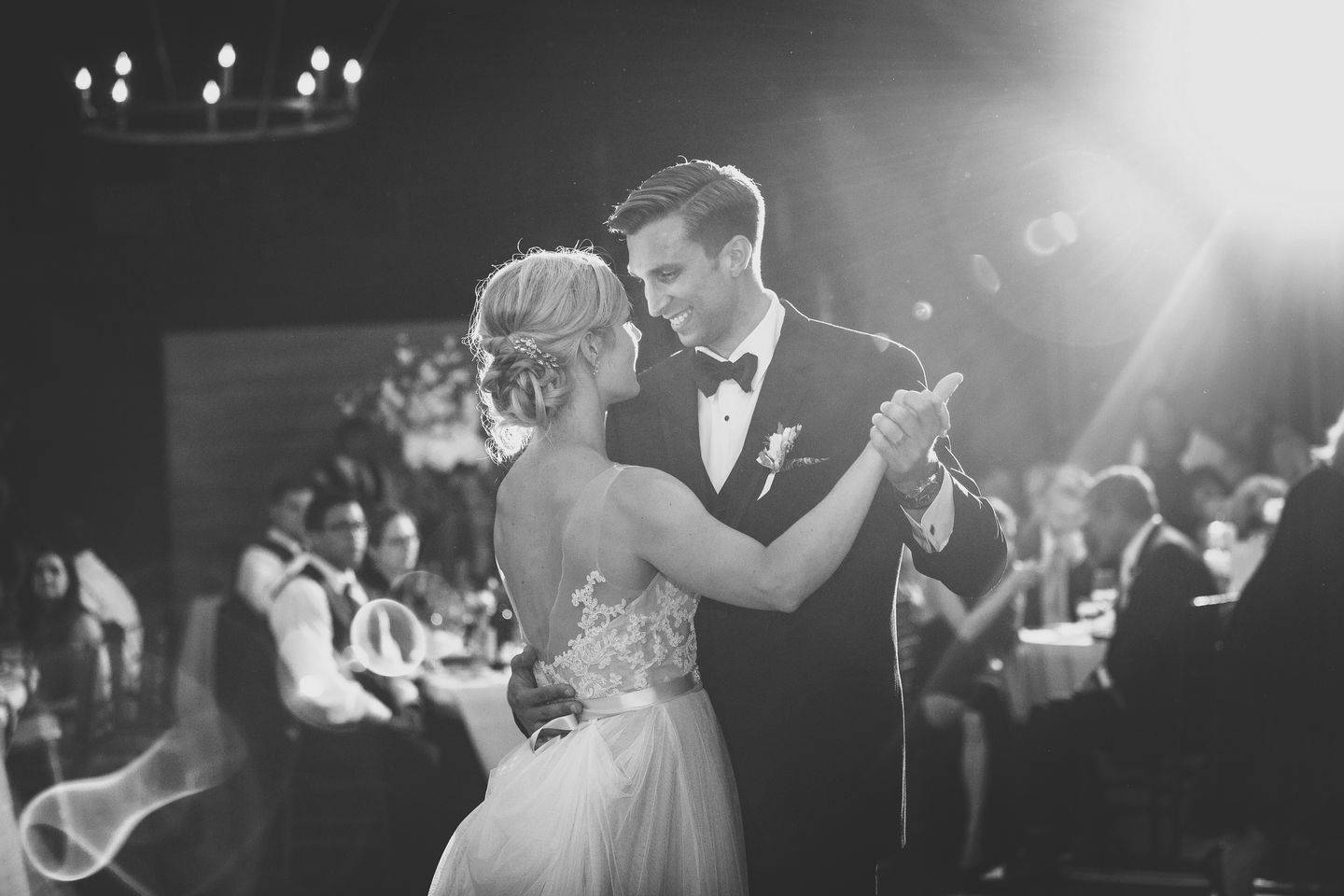a black and white photo of a bride and groom looking at each other and smiling while they share their first dance. Guests seated at tables are blurred out in the background, dramatic cinematic lighting streams through the dark room 