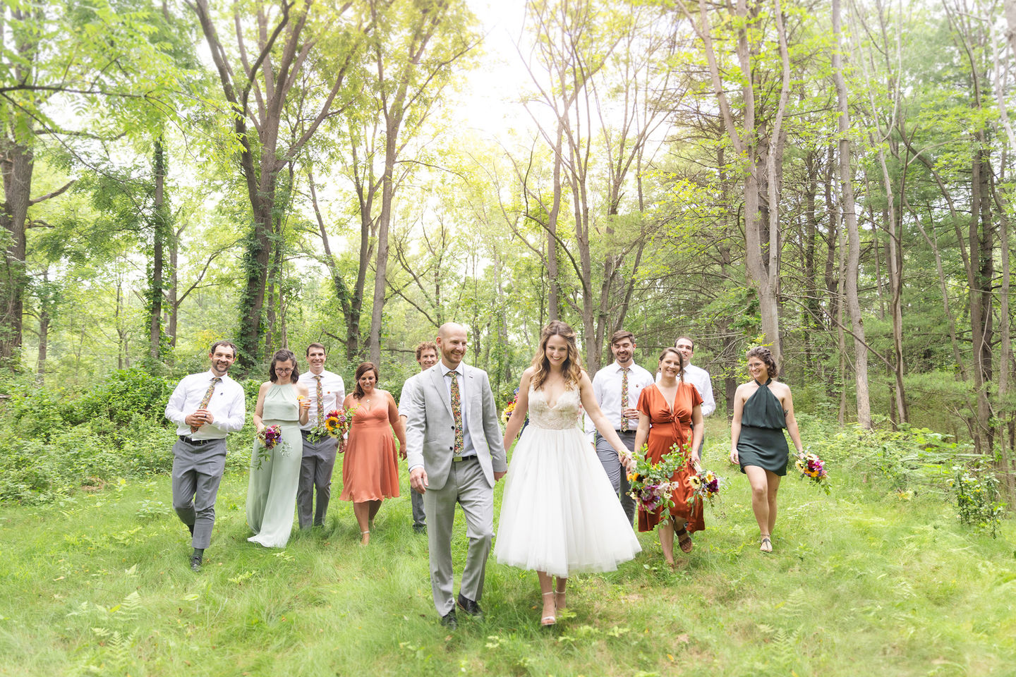 a bride and groom holding hands and walking through the woods with their colorfully dressed bridesmaids and groomsmen