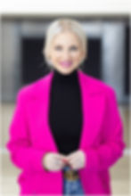 Smiling blonde woman in a bright pink blazer and black turtleneck, smiling confidently for a professional portrait.