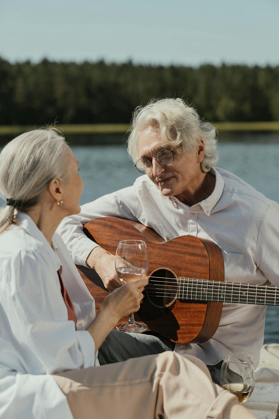 Middle-aged couple enjoying a romantic outdoor picnic with wine, as the man plays guitar for his partner. Both appear confident, relaxed, and deeply connected — reflecting the sense of security, emotional intimacy, and community support found in mature relationships over 50.