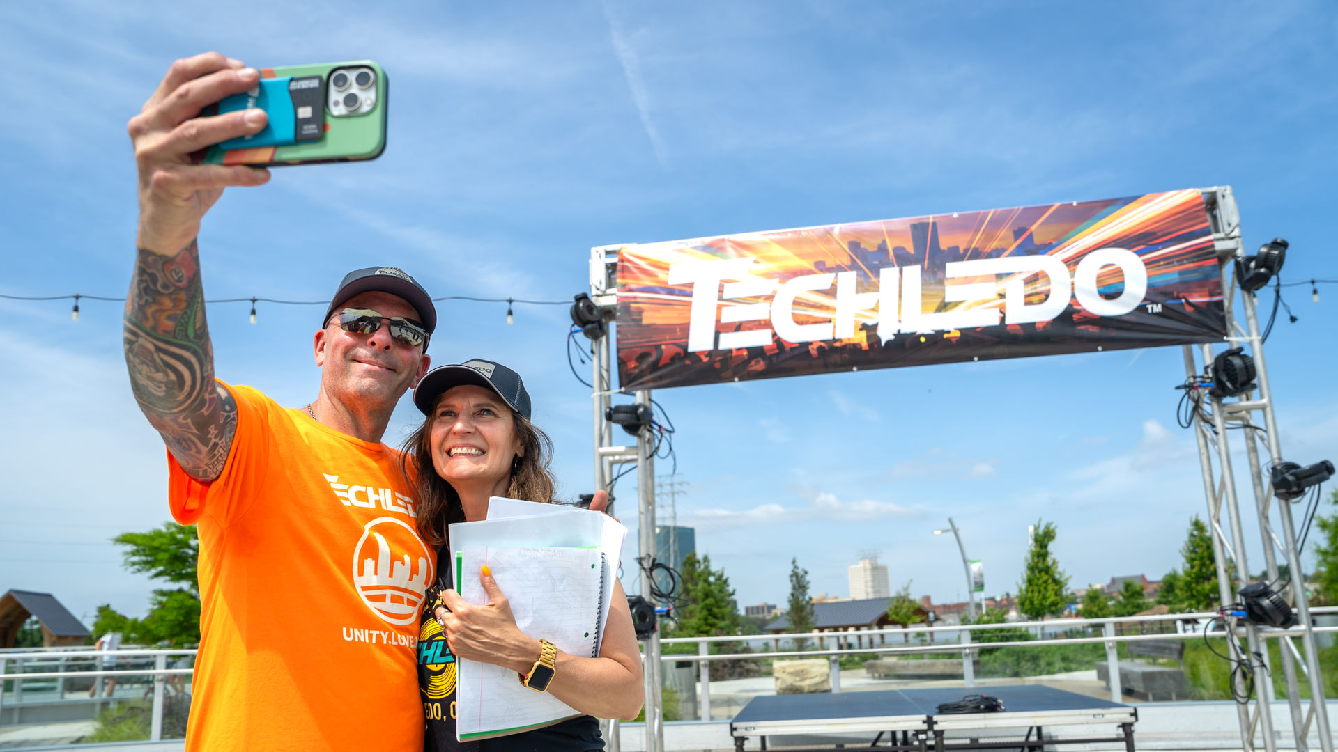 Two Tech Toledo team members smiling and taking a selfie in front of a Tech Toledo Unity Love Music stage at an outdoor event.