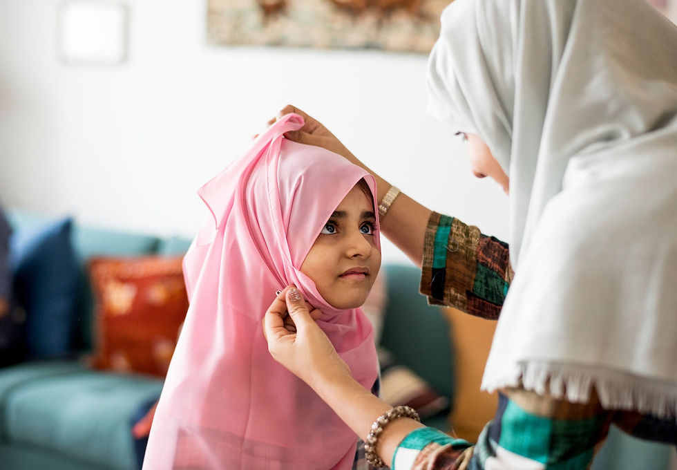 Mädchen mit rosafarbenem Hijab wird von einer Frau in einem hellen Raum angezogen. Sanft und fürsorglich, mit einem Sofa im Hintergrund.