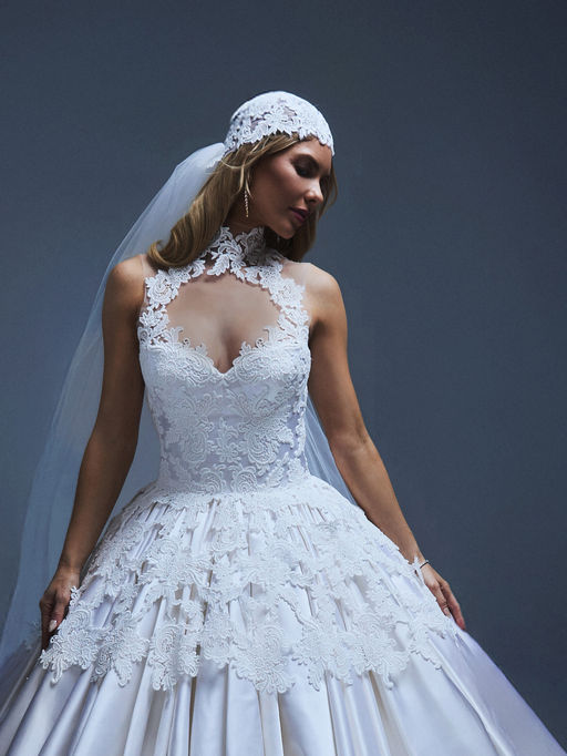 A vision of timeless grandeur, this regal ball gown is sculpted from luminous duchess satin and adorned with ornate guipure lace. The high collar and illusion tulle neckline frame the shoulders and neck with quiet elegance, while a row of covered buttons trails down the back, leading to a dramatic, sweeping train. Completing the look is a matching guipure lace headpiece and a long sweeping floor length veil.