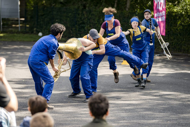 Mehrere Personen in blauen Arbeiterlatzhosen versuchen einer Person zu helfen, die offenbar mit dem Kopf in einer Tuba feststeckt.