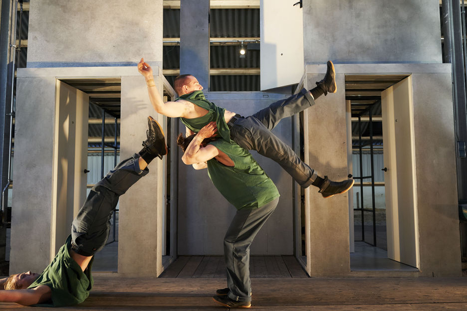 Drei Akrobaten bewegen sich vor einem Bühnenbild aus Betonwandelementen.