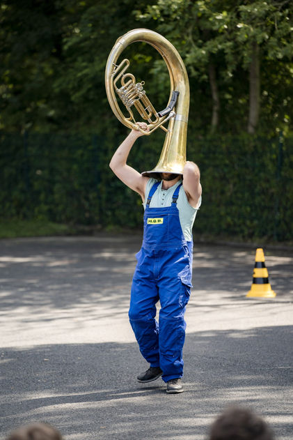 Eine Person in blauer Arbeiterlatzhose, die den Kopf in eine Tuba steckt.
